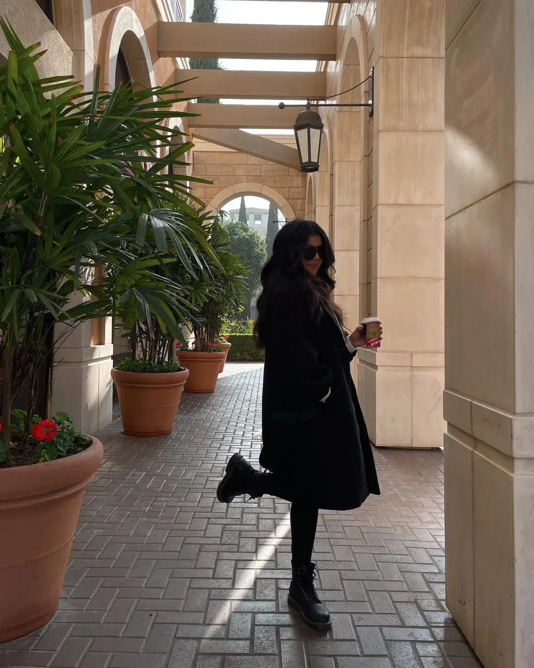 Valeria poses for a photo holding a coffee wearing a long, black coat and a boot.